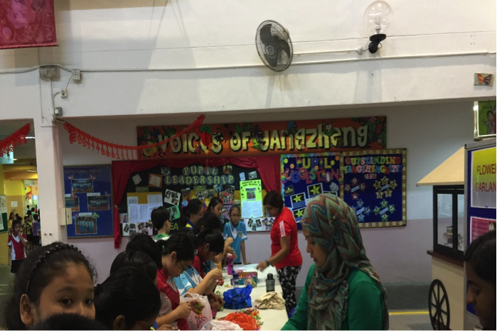 Children and adults gathered at tables in a school hall with a colorful "Voices of Jangzheng" banner on the wall.
